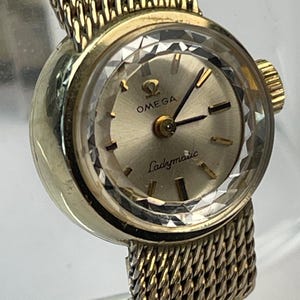 May include: A gold-tone Omega Ladymatic wristwatch featuring a round champagne-colored dial with gold indices, a faceted crystal edge, and a textured mesh metal bracelet. The watch face displays the Omega logo and the text Ladymatic.