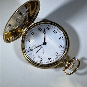 Könnte beinhalten: Eine goldene Taschenuhr mit weißem Zifferblatt und schwarzen Zeigern. Die Uhr hat ein kleines Sekundenzeiger-Zifferblatt und das Wort "ILLINOIS" ist auf dem Zifferblatt aufgedruckt.