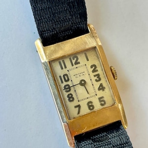 Könnte beinhalten: Eine goldfarbene Armbanduhr mit einem schwarzen Stoffband. Das Zifferblatt der Uhr ist weiß mit schwarzen römischen Ziffern für die Stunden und schwarzen arabischen Ziffern für die Minuten. Das Zifferblatt der Uhr enthält auch den Text "WALTHAM U.S.A."