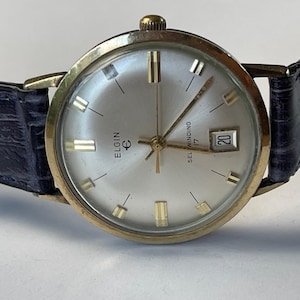 May include: Vintage Elgin self-winding wristwatch with a gold-tone case and a silver dial. The watch features gold-tone hands and markers, a date window, and a dark blue leather strap. The watch face has the word "ELGIN" and "SELF WINDING 17".