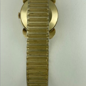 May include: A gold-toned wristwatch with a round face and a gold-toned metal band. The watch has a simple design and is in good condition.