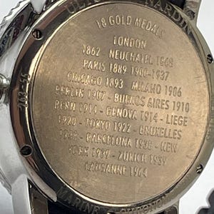May include: Close-up of a silver and black Ulysse Nardin Marine Chronometer watch. The watch back features engraved text: "18 Gold Medals" and lists cities and years, including London, Paris, and Tokyo. The watch has a black band.