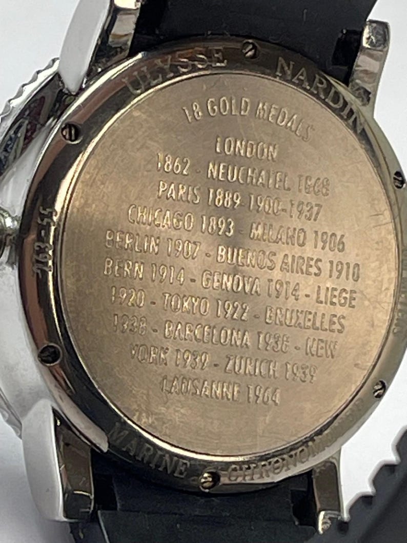 May include: Close-up of a silver and black Ulysse Nardin Marine Chronometer watch. The watch back features engraved text: "18 Gold Medals" and lists cities and years, including London, Paris, and Tokyo. The watch has a black band.