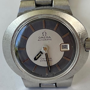 May include: A vintage Omega Genève Dynamic automatic wristwatch. The watch features a silver and dark gray dial with gold-toned hands and markers. The date window is at the 3 o'clock position. The watch has a stainless steel case and a textured bezel.