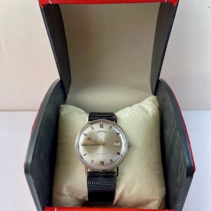 May include: A silver-toned wristwatch with a black leather band rests on a cream-colored pillow inside a red and black box. The watch face has silver hands and hour markers, with a date display at the 3 o'clock position. The box is open, revealing the watch.