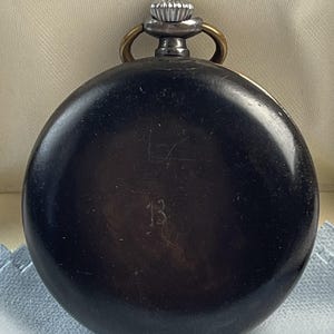 May include: A round, black pocket watch with a smooth, dark finish. The watch features a silver-toned winding crown at the top, flanked by two small brass-colored loops. The number 13 is engraved in the center of the black circular casing.