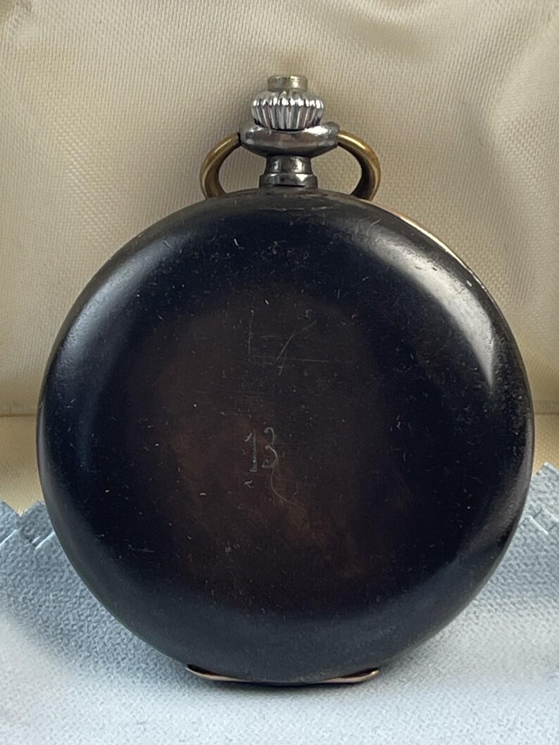 May include: A round, black pocket watch with a smooth, dark finish. The watch features a silver-toned winding crown at the top, flanked by two small brass-colored loops. The number 13 is engraved in the center of the black circular casing.