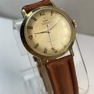May include: A vintage Waltham wristwatch with a gold-tone case and a brown leather band. The watch face is gold with black hands and hour markers. The watch reads "Waltham" and "Shock Resistant".