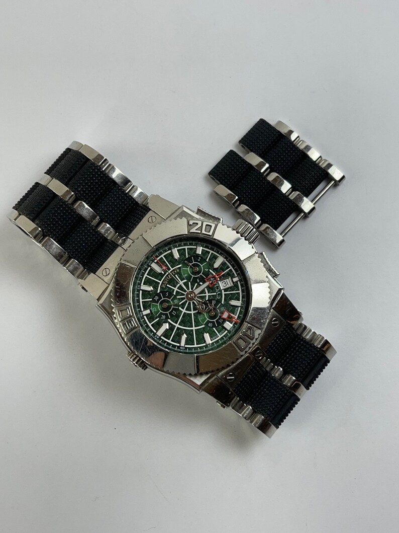May include: A silver wristwatch with a black rubber strap and a green face. The watch features a chronograph and a date window. The watch is displayed against a white background.