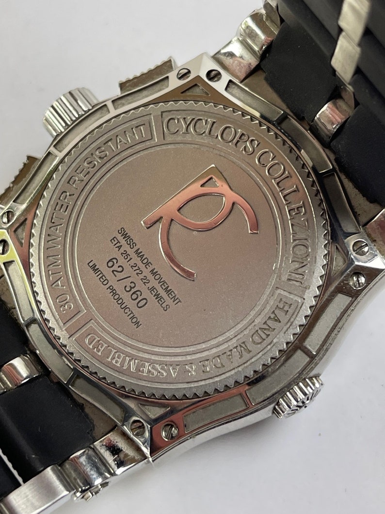 May include: A silver watch with a black rubber strap. The watch face is silver and has the text "Cyclops Collection Hand Made & Assembled 30 ATM Water Resistant" around the edge. The watch face also has the text "Swiss Made Movement ETA 251.272 22 Jewels 62/360 Limited Production" on it.