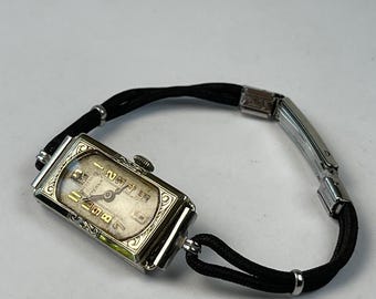 Può includere: Orologio da polso rettangolare vintage con cassa color argento e quadrante color crema. L'orologio presenta numeri e lancette neri, un cinturino in corda nero e una chiusura color argento. Un accessorio classico.