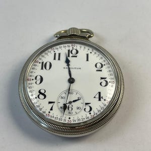 May include: A silver Hamilton pocket watch with a white face and black numerals. The watch features a smaller subdial and a textured bezel. The brand name "HAMILTON" is printed on the face.
