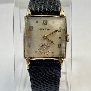 Op de afbeelding: Vintage Bulova horloge met een rechthoekige goudkleurige kast en een zwarte leren band. De crème-kleurige wijzerplaat heeft gouden wijzers en markeringen. De merknaam Bulova is zichtbaar.