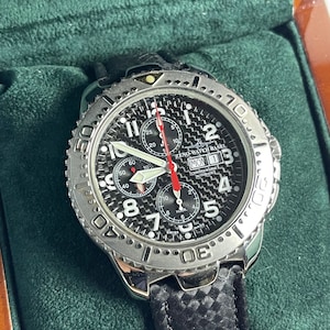 Può includere: Un orologio da polso cronografo Zeno-Watch Basel nero e argento con cinturino in pelle nera. Il quadrante dell'orologio presenta un motivo in fibra di carbonio e marcature bianche. L'orologio ha una finestra della data e una funzione cronografo.