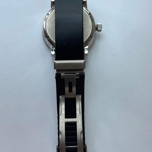 May include: A silver-toned wristwatch with a black band. The watch face is visible, and the band is open, revealing the clasp. The watch has a round face and a textured bezel.