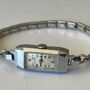 May include: A silver wristwatch with a rectangular face and a silver metal band. The watch face has black Roman numeral hour markers and a white background. The watch face also has the text "Rostorf Spezial" printed on it.