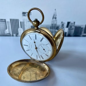 Puede incluir: Un reloj de bolsillo de oro con una esfera blanca y marcadores de hora romanos negros. El reloj está abierto y la tapa trasera está colocada a su lado.