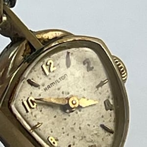 May include: A gold-tone Hamilton wristwatch with a triangular case and a textured off-white dial. The watch face features gold-tone hour markers at the 12, 10, 8, 6, 4, and 2 positions, with gold-tone hands and a small crown on the right side.