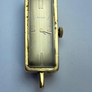 May include: A vintage Bulova wristwatch with a rectangular gold-tone case. The watch face is a light gold color with black hands and hour markers. The word "Bulova" is printed on the watch face. The watch has a gold-tone crown and a small gold-tone bar at the bottom.