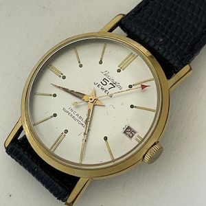 May include: A gold-toned wristwatch with a white face and black leather band. The watch face has black Roman numeral hour markers, black stick minute markers, and a date window at the 3 o'clock position. The watch face also features the text "Lexington 517 Jewels Incabloc Superautomatic".