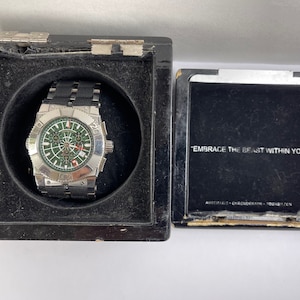 May include: A silver wristwatch with a black strap and a green face. The watch is in a black box with the words "Embrace the Beast Within You" on the lid.
