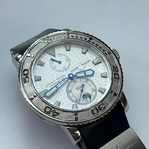 May include: A silver Ulysse Nardin watch with a white face, blue hands, and a black band. The watch face has multiple sub-dials and the text "OFFICIALLY CERTIFIED CHRONOMETER". The watch is a luxury timepiece.