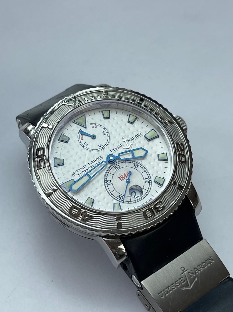 May include: A silver Ulysse Nardin watch with a white face, blue hands, and a black band. The watch face has multiple sub-dials and the text "OFFICIALLY CERTIFIED CHRONOMETER". The watch is a luxury timepiece.
