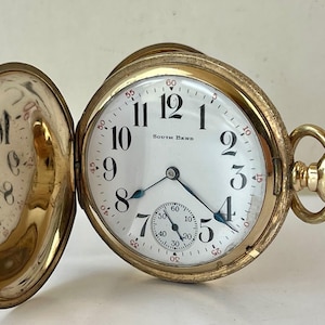 Könnte beinhalten: Eine goldfarbene Taschenuhr mit offenem Zifferblatt, die ein weißes Zifferblatt mit schwarzen Ziffern und Zeigern zeigt. Auf dem Zifferblatt steht "South Bend". Ein kleineres Hilfszifferblatt befindet sich unten. Die Uhr ist geöffnet.