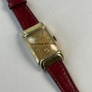 Puede incluir: Un reloj de pulsera Bulova vintage con una caja rectangular dorada y una esfera color crema. El reloj presenta agujas e indicadores de hora dorados, y la marca es visible. Tiene una correa de cuero roja.