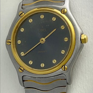Op de afbeelding: Een zilver- en goudkleurig Ebel quartz horloge met een donkergrijze wijzerplaat. Het horloge heeft gouden wijzers en uurmarkeringen, en de band is een zilver- en goudkleurige schakelstijl. Het horloge heeft een ronde wijzerplaat en een gouden kroon.
