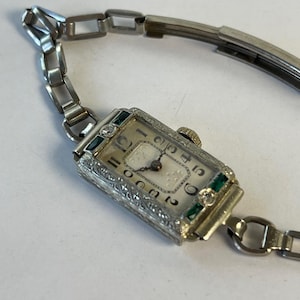 May include: A vintage silver wristwatch with a rectangular face. The watch features a white dial with black numerals and hands, and is accented with green and clear gemstones. The watch band is made of silver links.