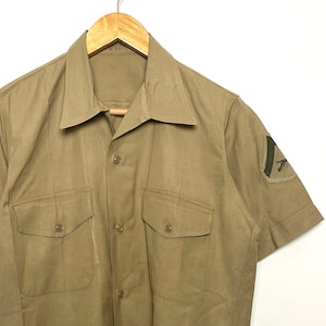 May include: A tan short-sleeved shirt with two pockets and a button-up front. The shirt has a patch on each sleeve with a green symbol.