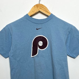 May include: A light blue t-shirt featuring a maroon Philadelphia Phillies logo and a white Nike swoosh.