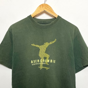 May include: A green t-shirt featuring a yellow graphic of a person skateboarding. The graphic is made up of the word "Abercrombie" and the words "skateboarding".