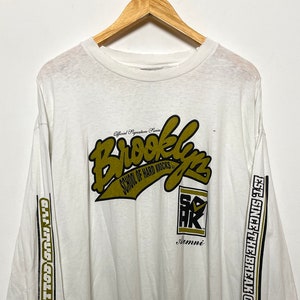 Könnte beinhalten: Weißes Langarm-T-Shirt mit einer goldenen Grafik, auf der "Brooklyn School of Hard Knocks" steht, und einer schwarzen und goldenen Grafik, auf der "S.O.H.K. Alumni" steht. Die Ärmel haben schwarze und goldene Streifen mit dem Text "EST. SINCE THE BREAK OF DAWN!" und "GARBAGE COLLECTOR!"