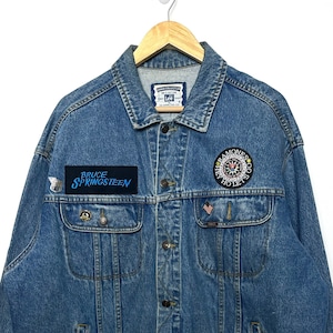 Puede incluir: Una chaqueta vaquera azul con varios parches, incluyendo un parche de "Bruce Springsteen" y un parche de "Ramones". La chaqueta tiene cierre de botones y dos bolsillos delanteros.