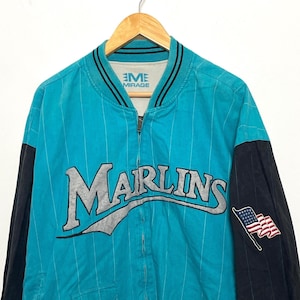 May include: A teal blue and black baseball jacket with a white "Marlins" logo and a small American flag patch on the sleeve. The jacket has a zipper closure and a striped pattern.