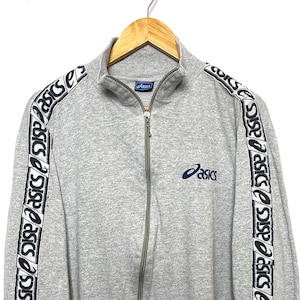 May include: A gray zip-up jacket with long sleeves. The sleeves have a white and blue stripe with the word "asics" repeated along the stripe. The jacket has a small "asics" logo on the chest.