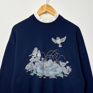 以下が含まれることがあります： 白のモックタートルネックカラーが付いたネイビーブルーのスウェットシャツ。前面には、ピンクの花のある噴水の近くにいる鳥のグラフィックが描かれています。鳥と噴水は銀色のグリッターで装飾されています。