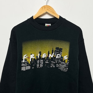 Peut inclure: Sweat-shirt noir avec un motif jaune et noir représentant la skyline de New York et le mot "FRIENDS" dans une police colorée.