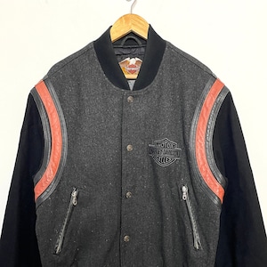 May include: A gray wool varsity jacket with black sleeves and red and black leather accents. The jacket has a snap closure and a Harley Davidson logo on the front.