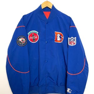Op de afbeelding: Blauwe Starter-jas met oranje details en meerdere patches, waaronder een Coca-Cola-logo, een Denver Broncos-logo en een NFL-patch. De jas heeft een knoopsluiting en een rode voering.