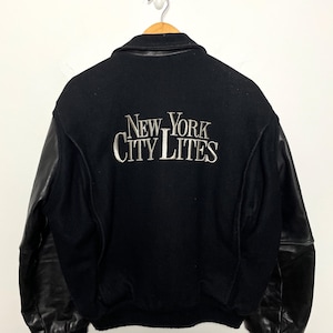 May include: Black varsity jacket with leather sleeves. The back of the jacket features the embroidered text "NEW YORK CITY LITES" in silver. The jacket is hanging on a wooden hanger against a white background.