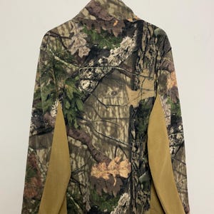 May include: A camouflage fleece jacket with a button-up collar and tan side panels. The jacket features a woodland pattern with shades of green, brown, and beige. The back of the jacket is visible, hanging from a hanger.