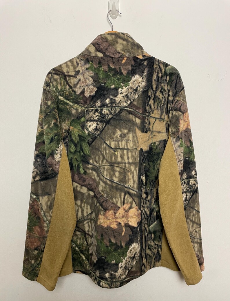 May include: A camouflage fleece jacket with a button-up collar and tan side panels. The jacket features a woodland pattern with shades of green, brown, and beige. The back of the jacket is visible, hanging from a hanger.
