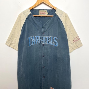 May include: A blue button-down baseball jersey with cream-colored raglan sleeves featuring thin pinstripes. The word TARHEELS is embroidered in light blue across the chest. A Mirage First String label is visible at the neck and on the sleeve.