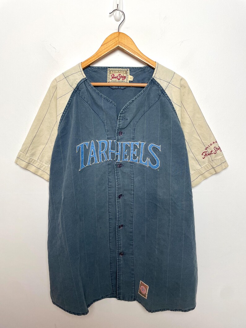 May include: A blue button-down baseball jersey with cream-colored raglan sleeves featuring thin pinstripes. The word TARHEELS is embroidered in light blue across the chest. A Mirage First String label is visible at the neck and on the sleeve.