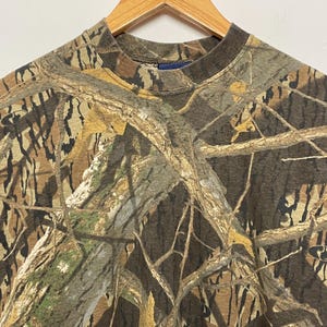 May include: A camouflage pattern t-shirt with a crew neck, hanging on a wooden hanger. The shirt features a brown, green, and tan woodland camouflage design. The fabric appears to be a medium-weight material.