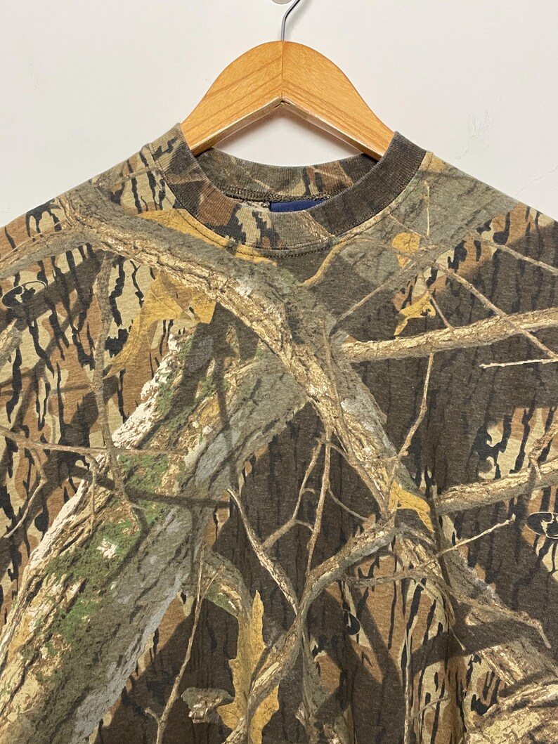 May include: A camouflage pattern t-shirt with a crew neck, hanging on a wooden hanger. The shirt features a brown, green, and tan woodland camouflage design. The fabric appears to be a medium-weight material.