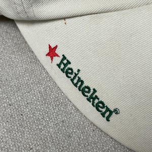 May include: A cream-colored baseball cap featuring an embroidered red star and the green text Heineken on the side of the brim. The fabric has a visible twill weave texture, and the stitching is consistent with standard cap construction.